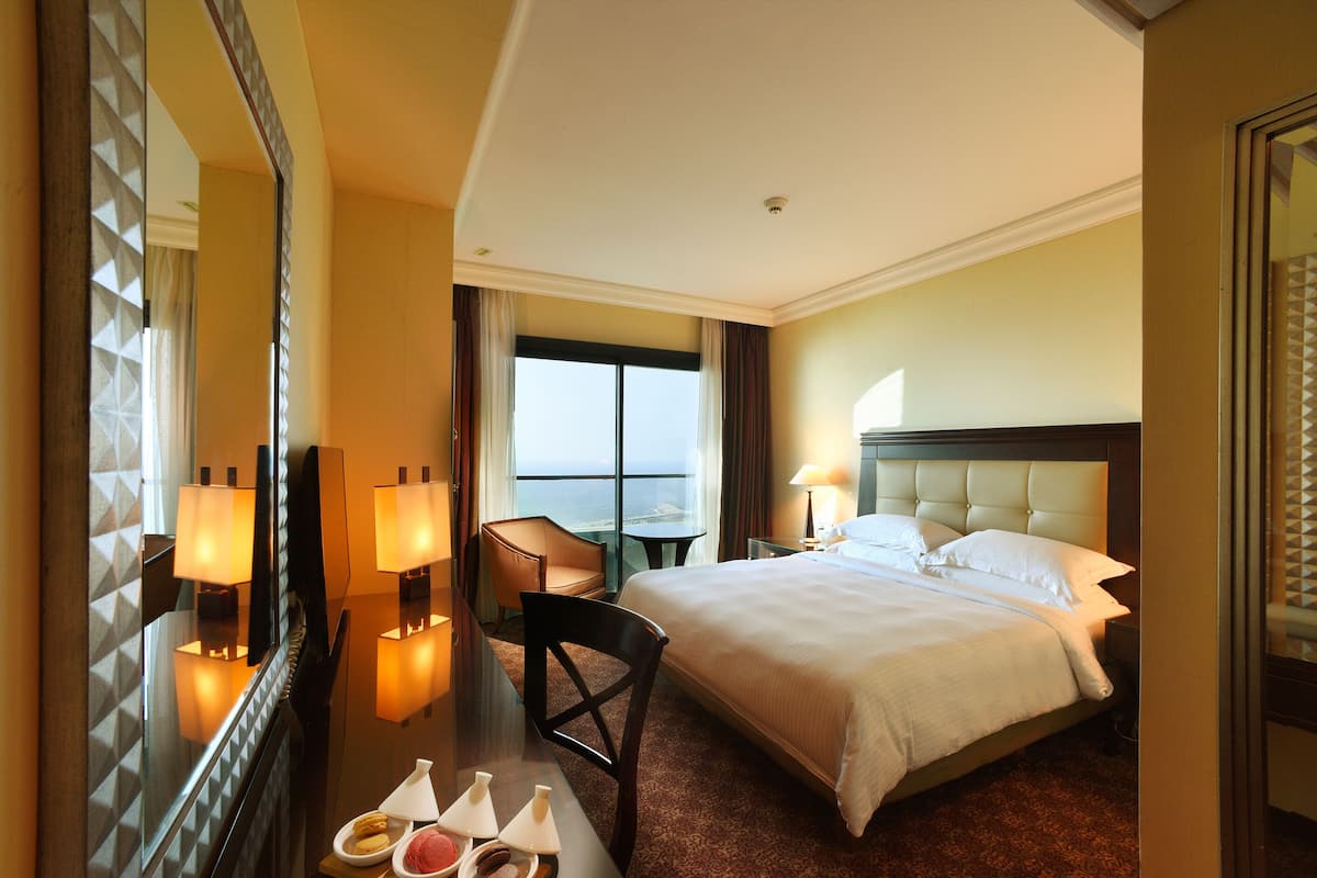 Habitación de hotel con una cama grande, un escritorio con una silla, un espejo y vista al mar.