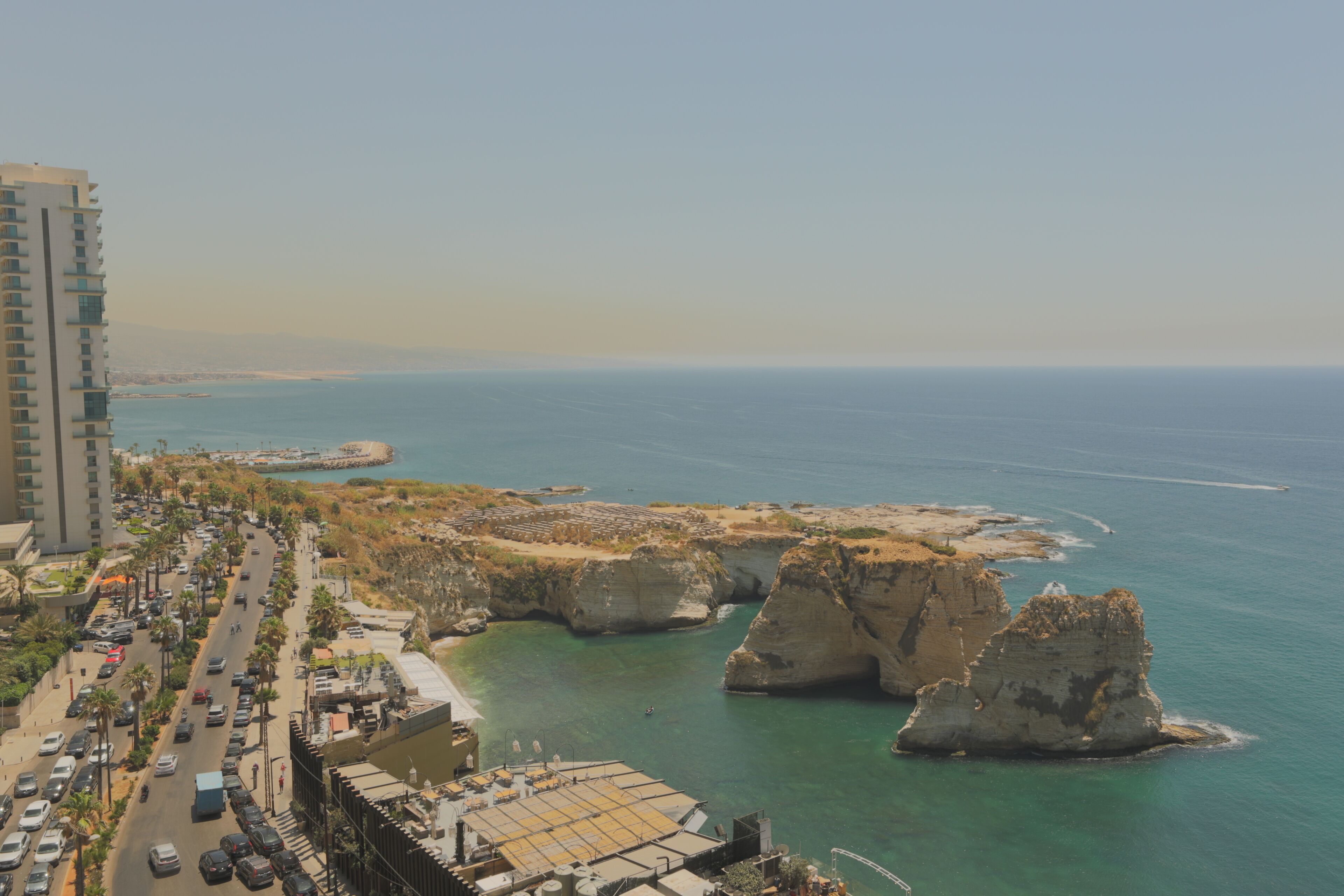 Un paysage urbain côtier avec un bâtiment imposant, une route bordée de voitures et des falaises rocheuses au bord de la mer.