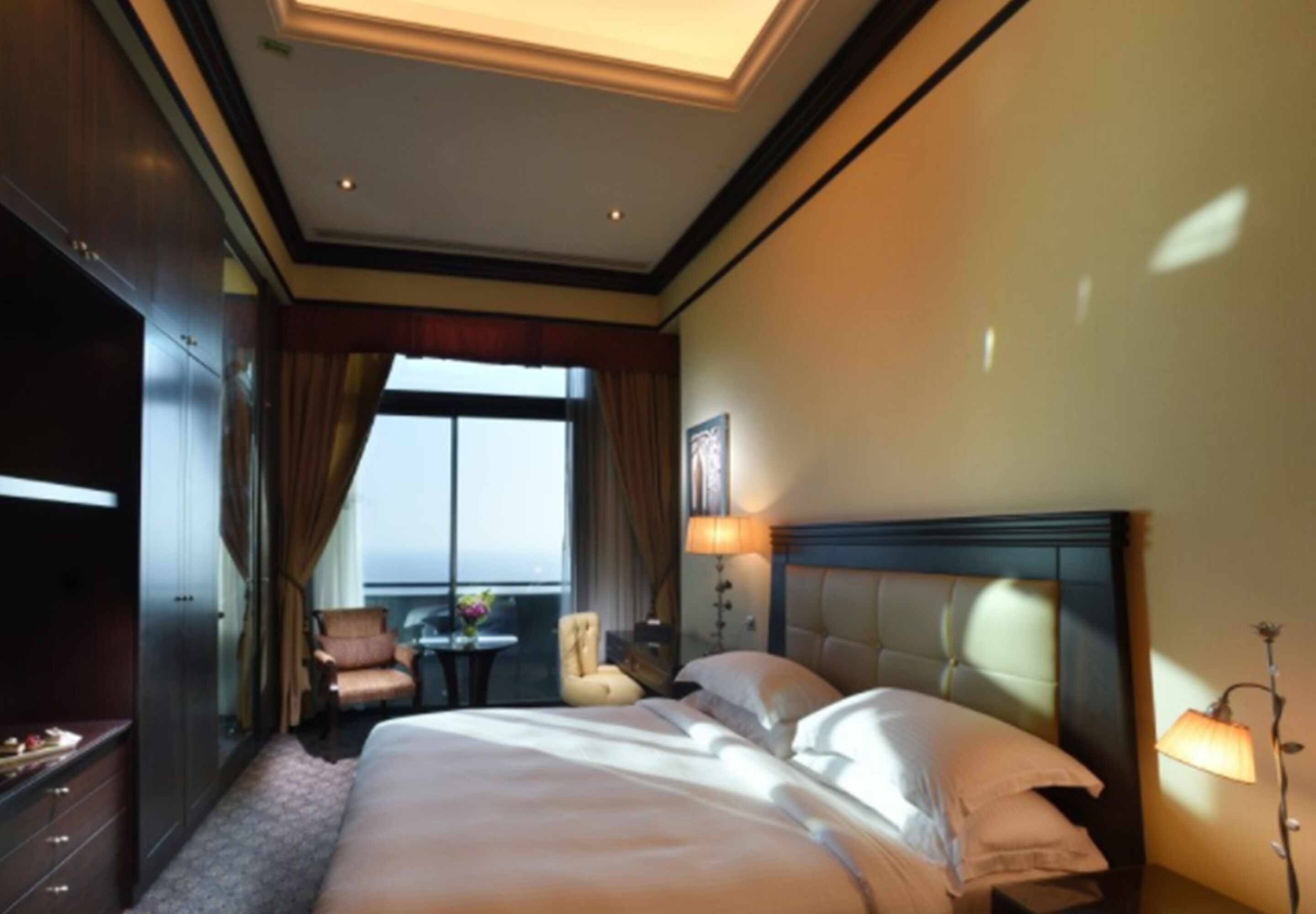 A hotel room with a large bed, a chair, a small table with a vase, and a view of the outdoors through a window.