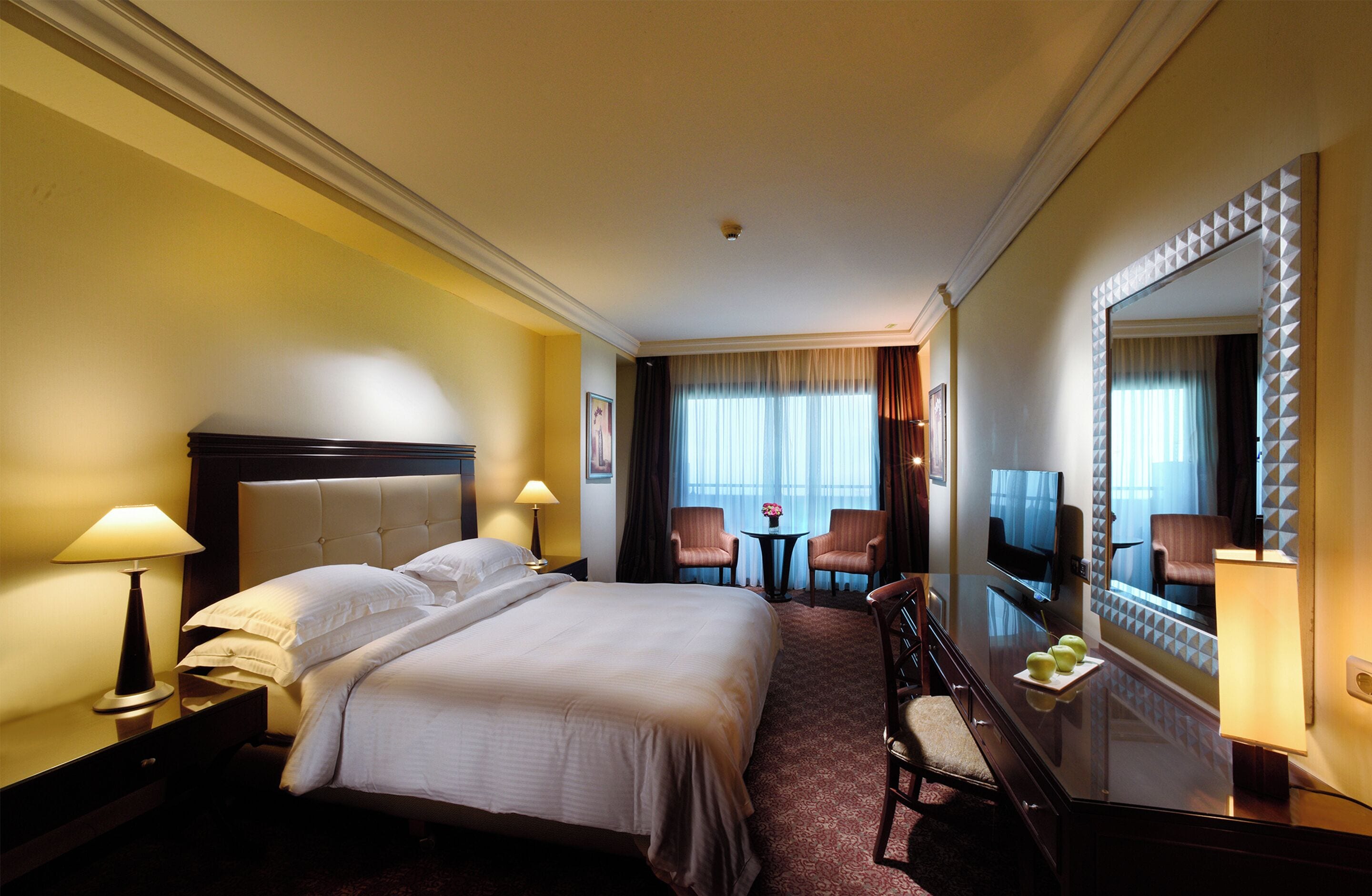 Une chambre d’hôtel avec un grand lit, deux fauteuils, une petite table, un téléviseur et un miroir orné d’un motif géométrique.