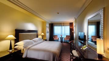 Une chambre d’hôtel avec un grand lit, deux fauteuils, une petite table, un téléviseur et un miroir orné d’un motif géométrique.