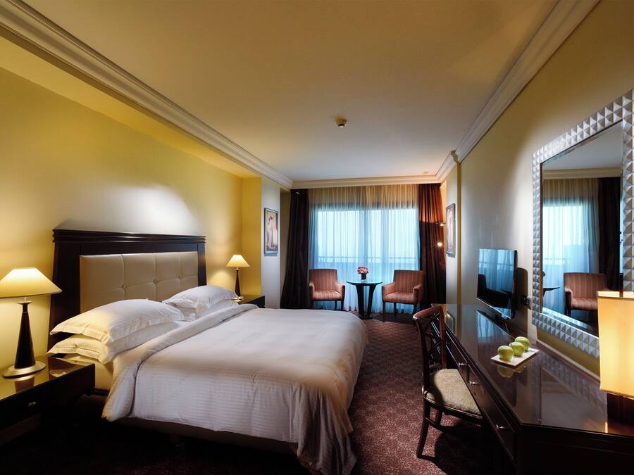 Une chambre d’hôtel avec un grand lit, deux fauteuils, une petite table, un téléviseur et un miroir orné d’un motif géométrique.