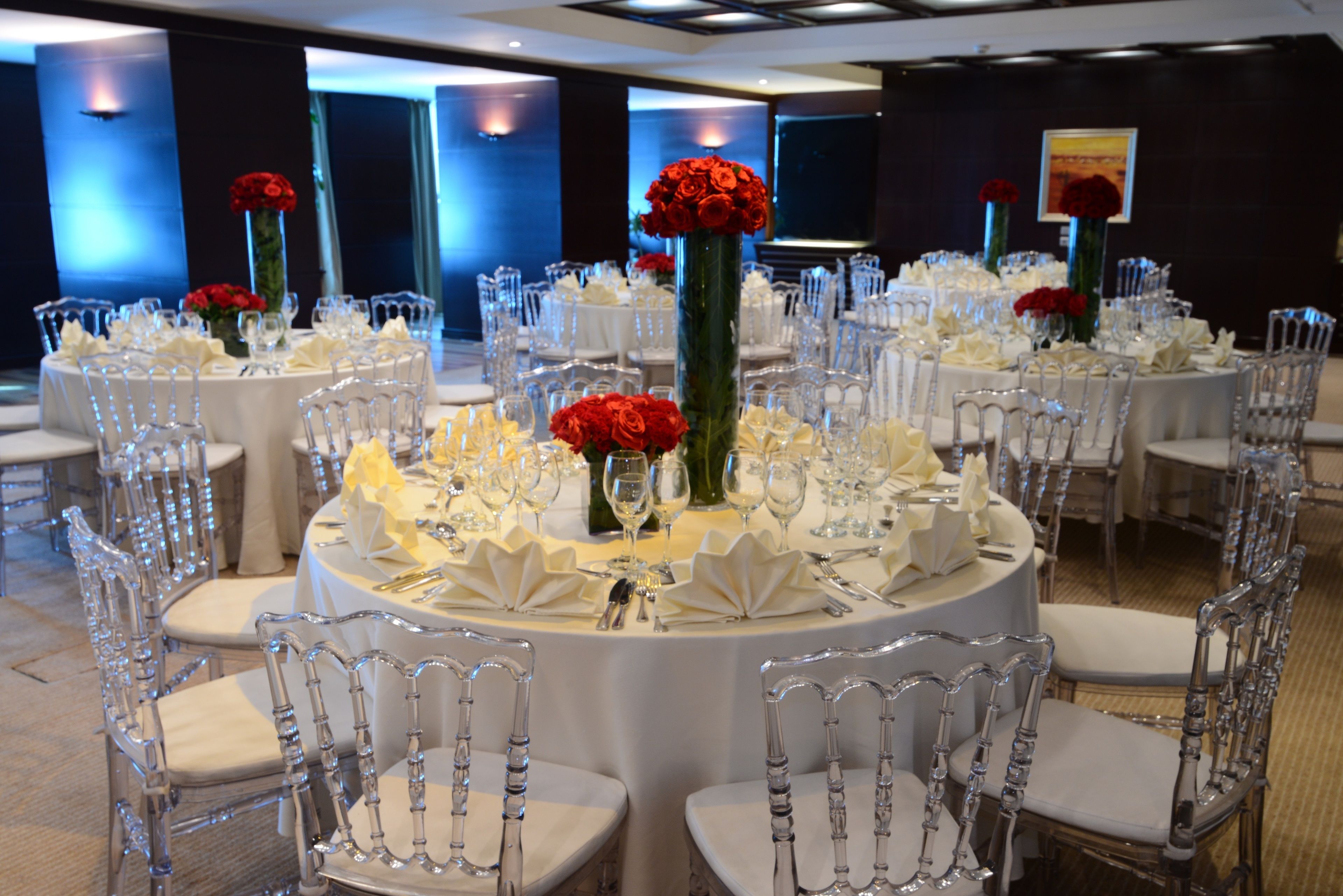 A banquet hall with round tables, clear chairs, and red roses in vases.