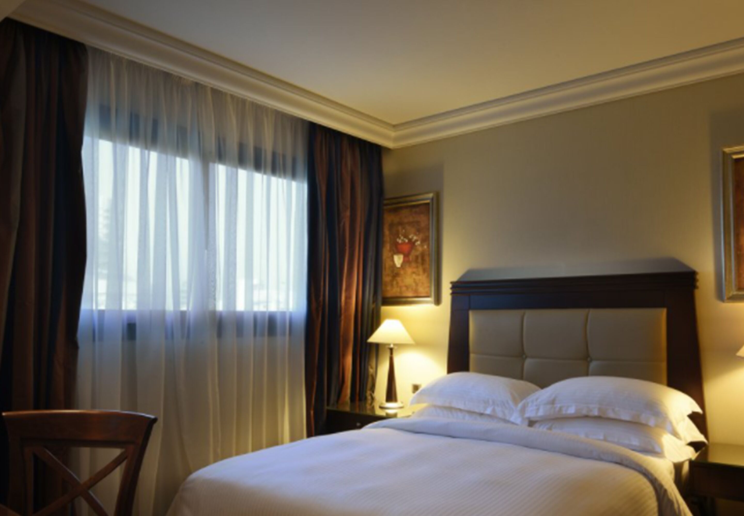 A hotel room with a large bed, a chair, a nightstand with a lamp, and a painting on the wall.