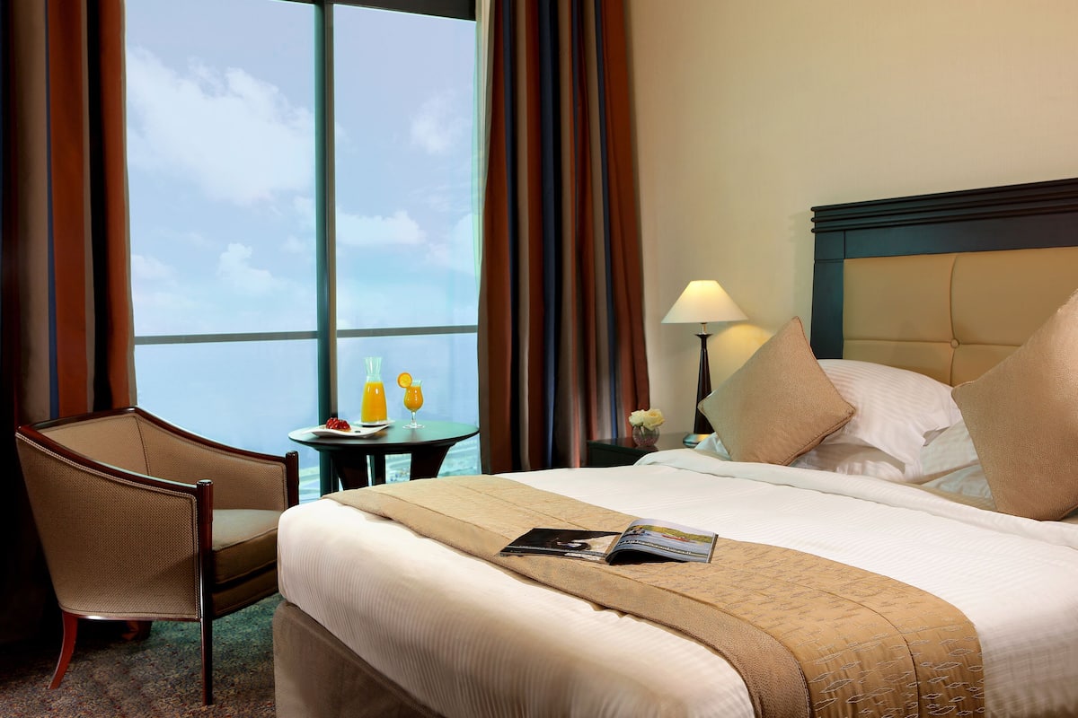Habitación de hotel con una cama grande, una silla, una mesita con bebidas y vista al cielo a través de la ventana.