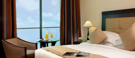 A hotel room with a large bed, a chair, a small table with drinks, and a view of the sky through the window.