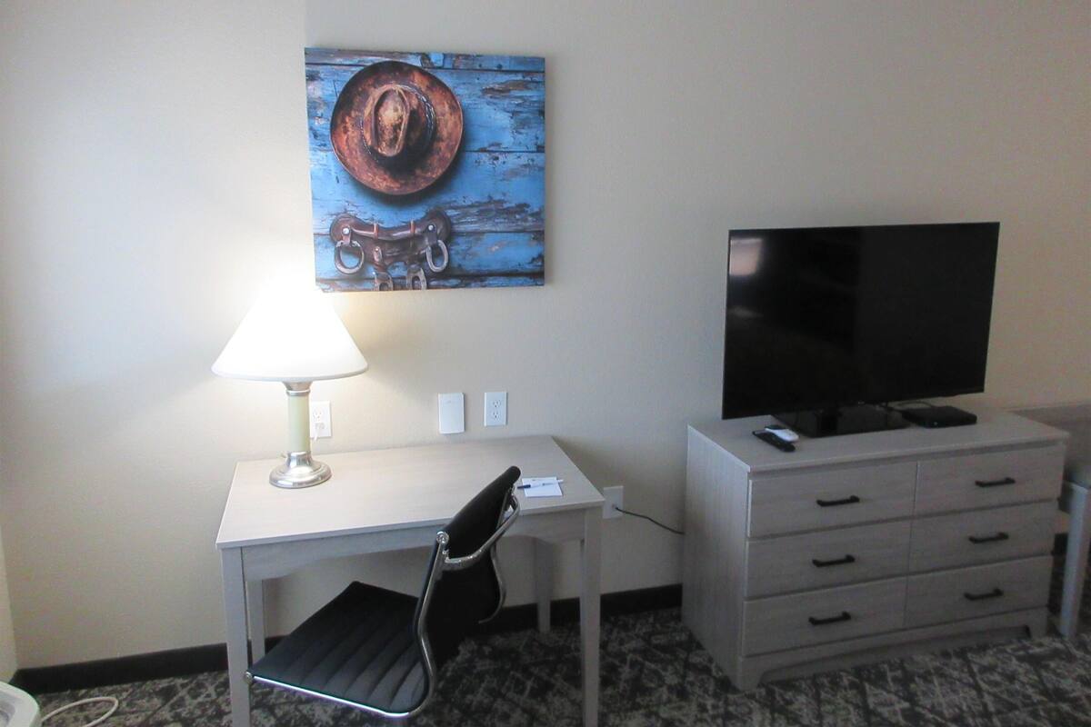 A hotel room with a desk, chair, TV, and a painting on the wall.