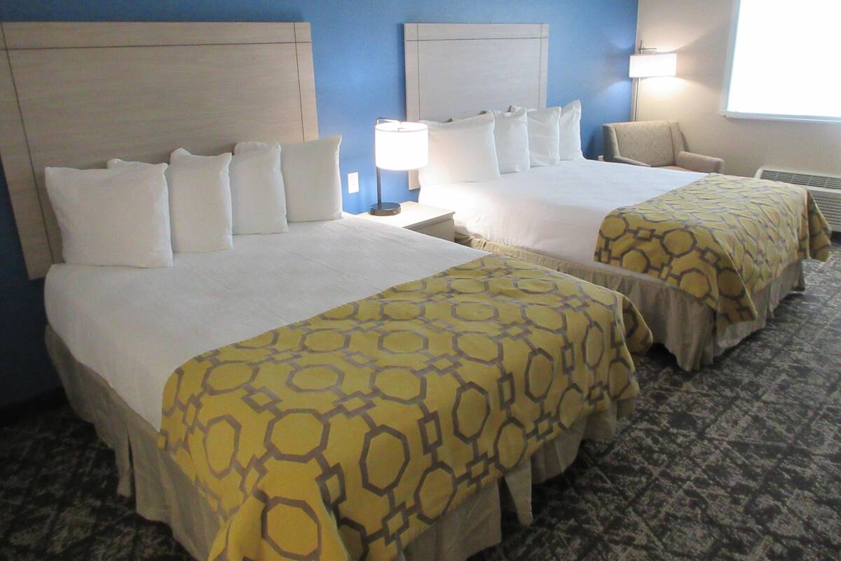 A hotel room with two beds, each with white bedding and a yellow patterned bedspread. There is a nightstand with a lamp, a chair, and a window with blinds.