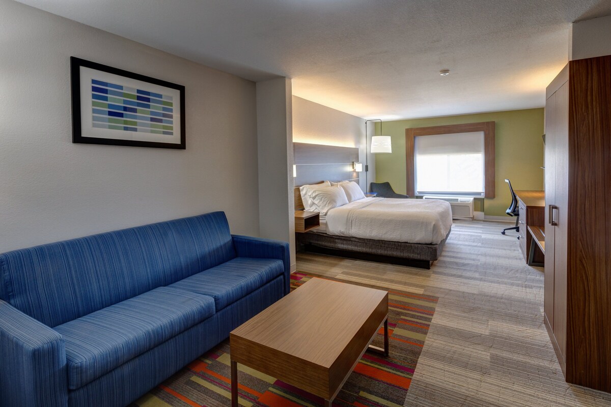 Une chambre d’hôtel avec un canapé bleu, une table basse en bois, un lit recouvert d’une couette blanche, une table de chevet, une lampe et une fenêtre avec des stores.