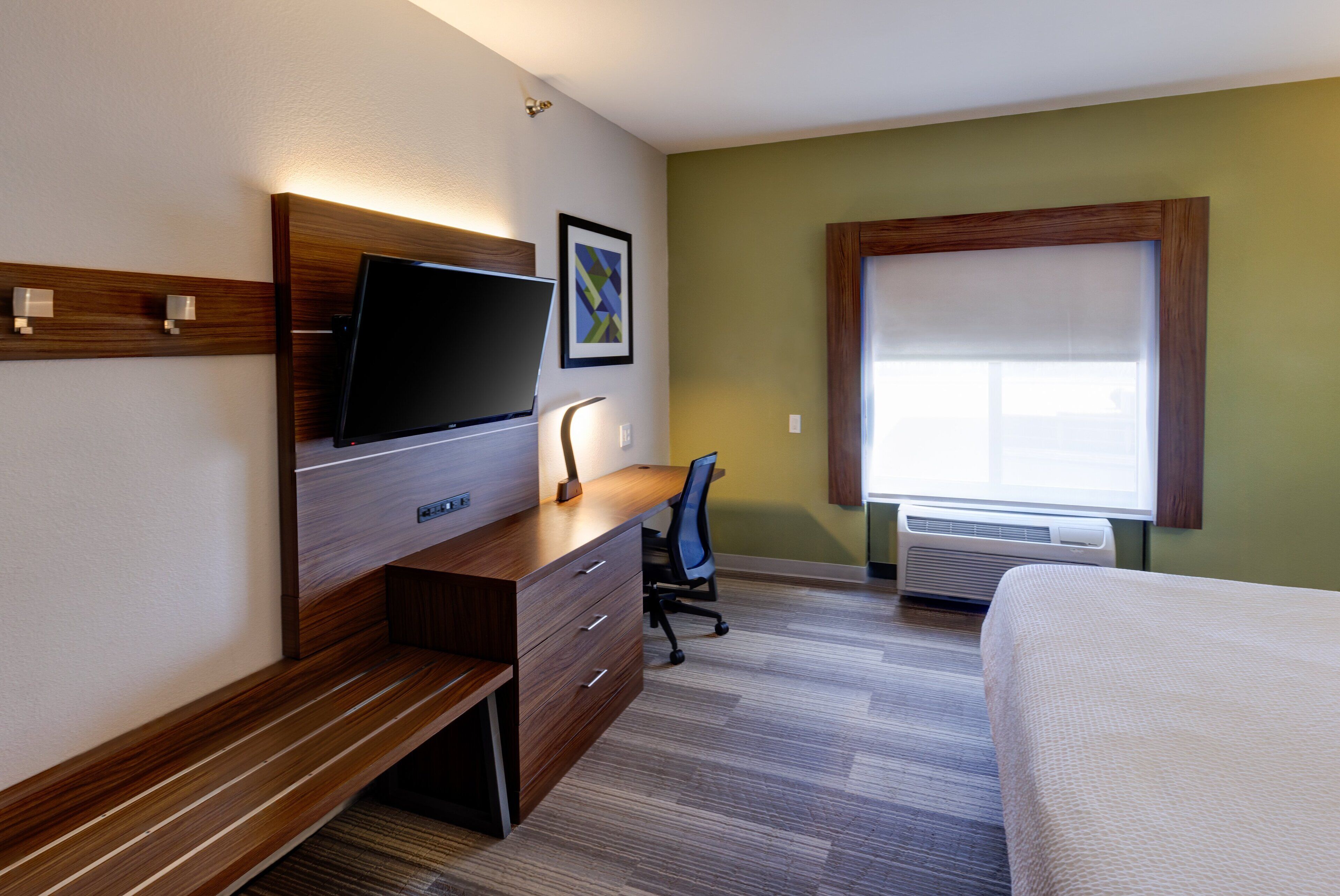A hotel room with a wooden desk, a flat-screen TV, a chair, a bed, and a window with blinds.