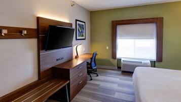A hotel room with a wooden desk, a flat-screen TV, a chair, a bed, and a window with blinds.