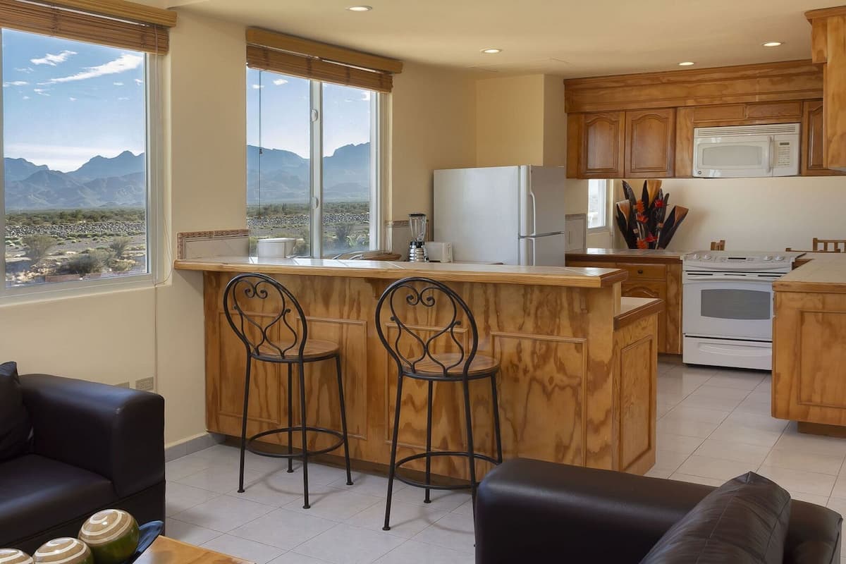 Una cocina con armarios de madera, un refrigerador blanco, un microondas y una estufa. Hay dos taburetes de bar y una ventana con vistas a montañas.
