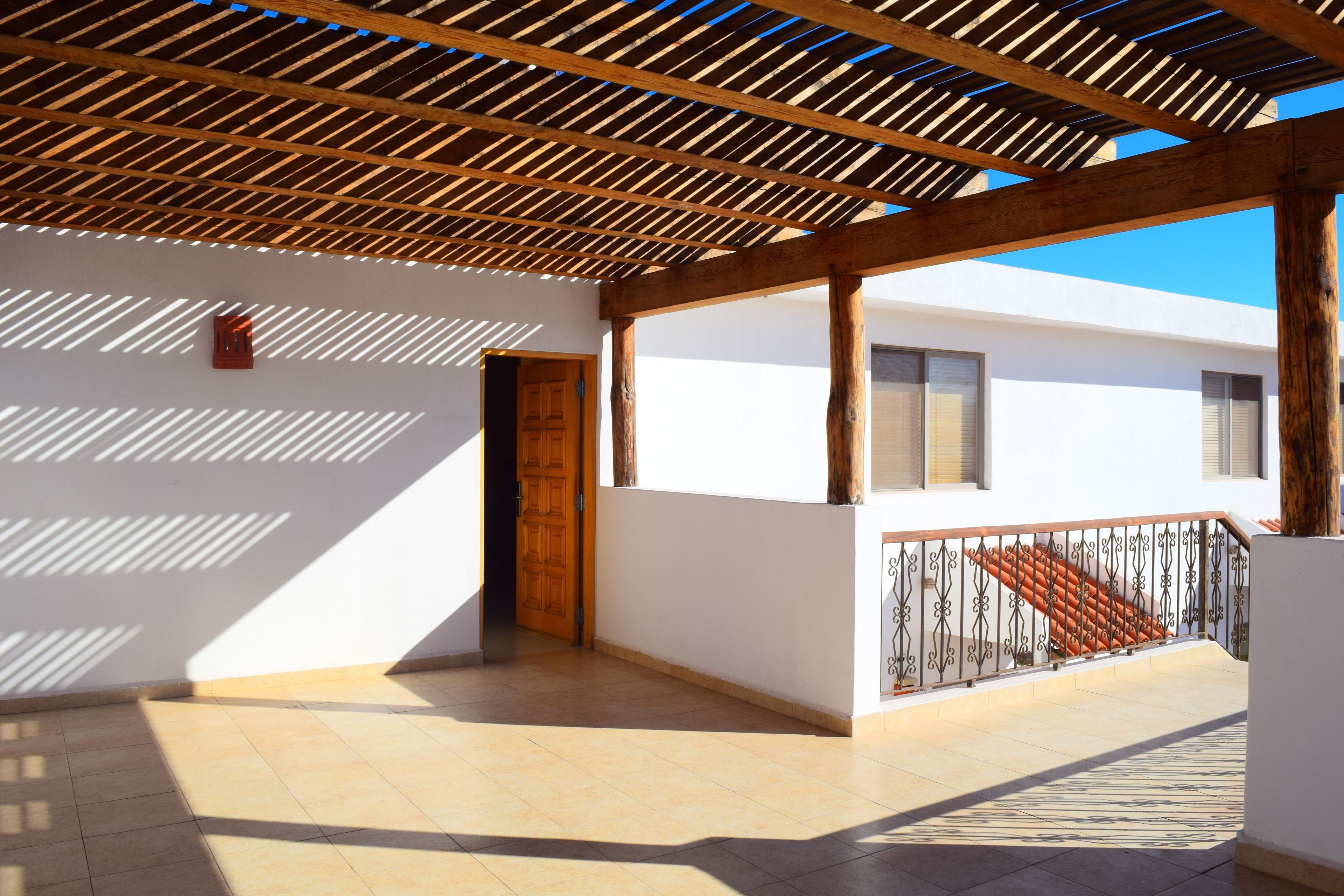 A covered outdoor area with a wooden ceiling, a tiled floor, and a white building with a door and windows.