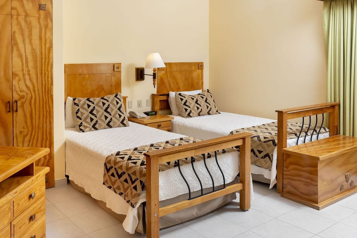 Habitación de hotel con dos camas de madera, una mesita de noche con lámpara, un aparador de madera y un armario.