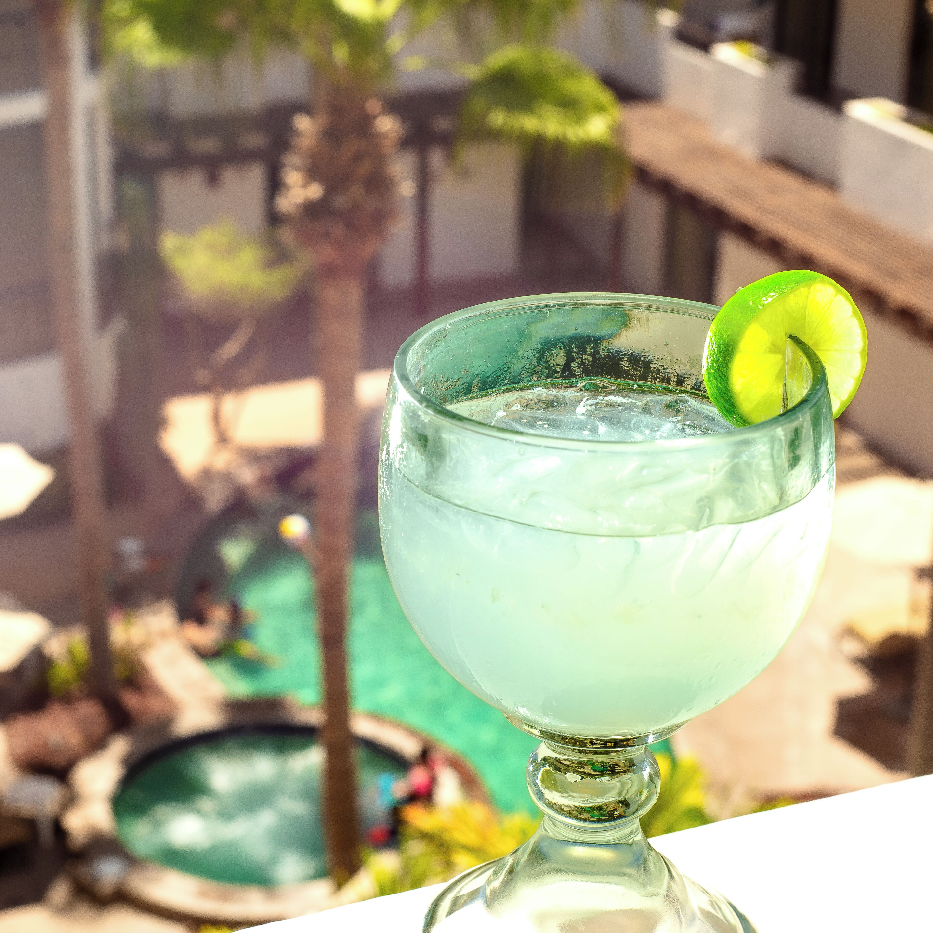 A cocktail glass with a lime garnish on a balcony overlooking a swimming pool.