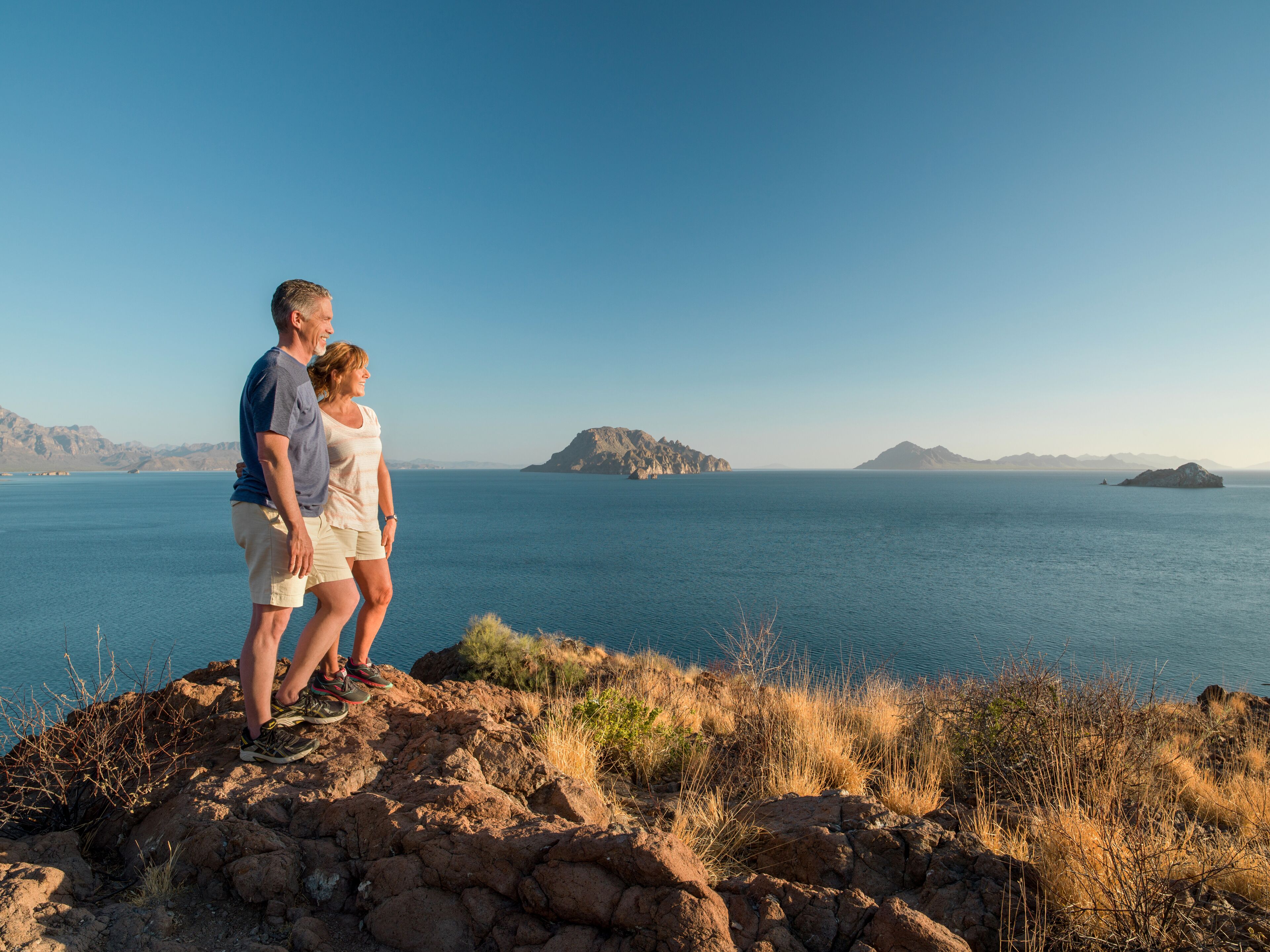 A couple standing on a rocky outcrop overlooking a calm sea with distant islands.