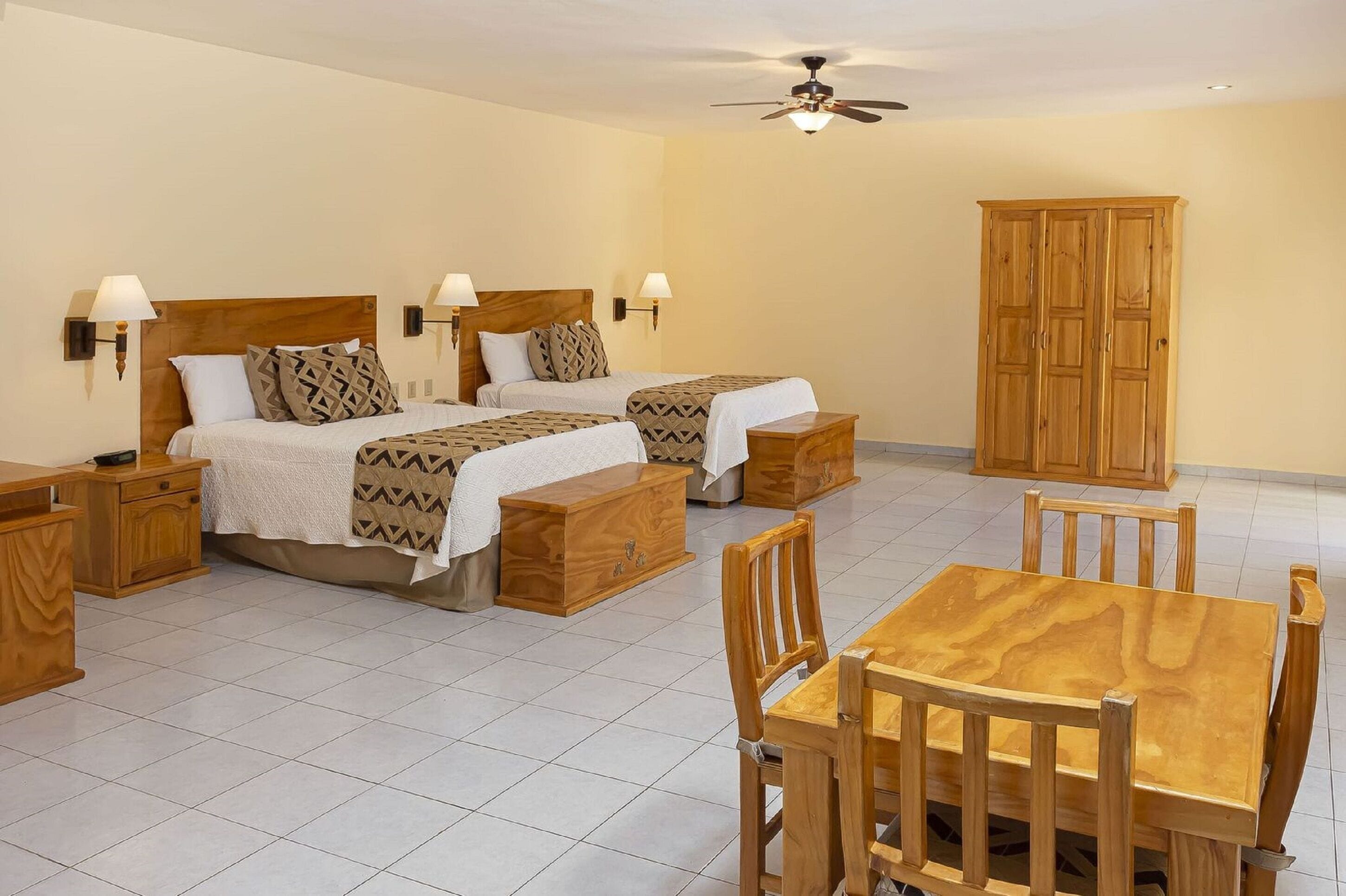 Une chambre d’hôtel avec deux lits, une armoire en bois, une table à manger avec des chaises et un ventilateur de plafond.