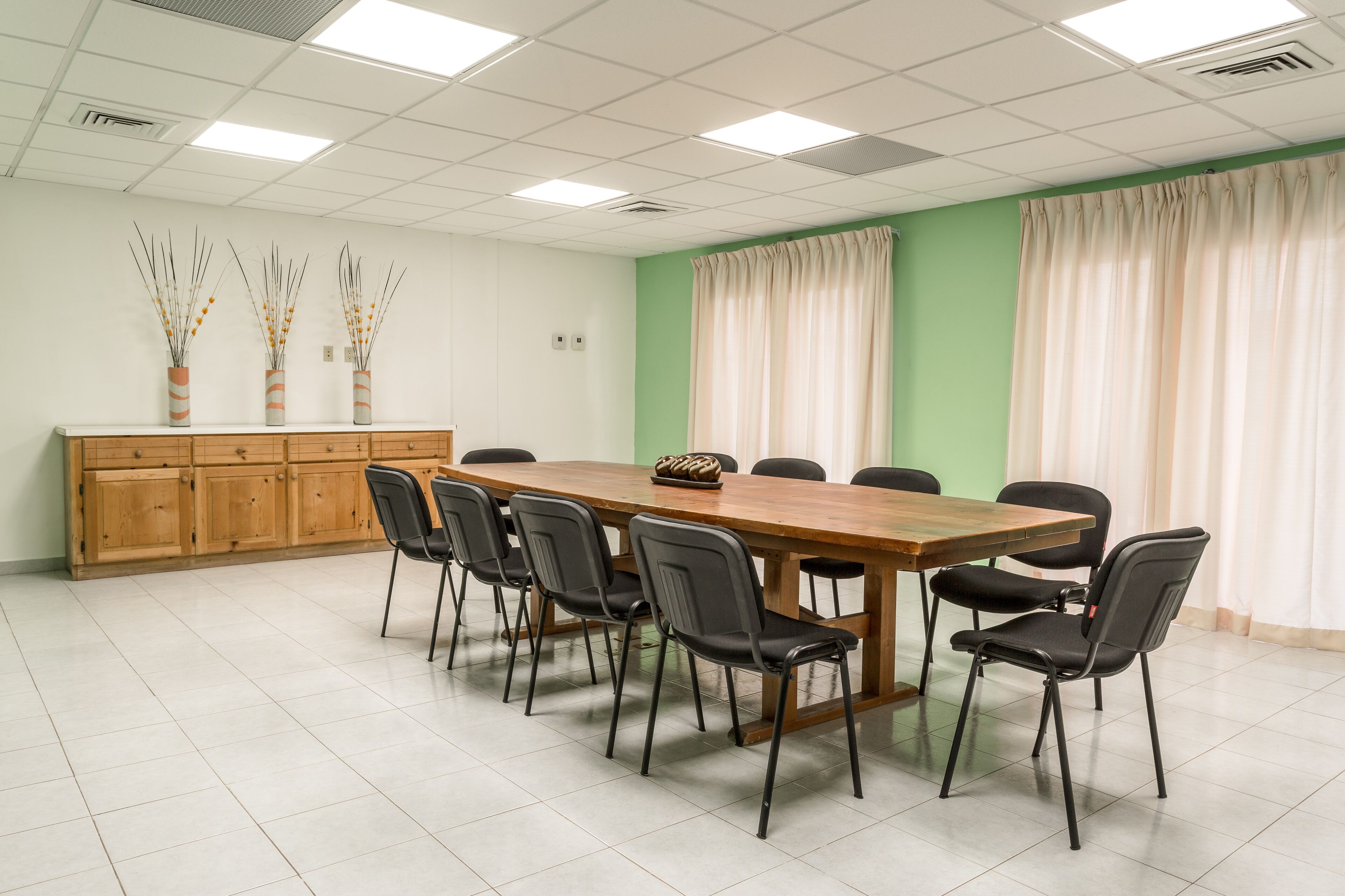 A conference room with a long wooden table, black chairs, a wooden cabinet, and three tall vases with dried plants.
