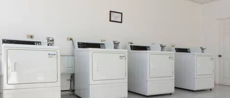 A row of white laundry machines in a clean, tiled room with a white door and a framed certificate on the wall.