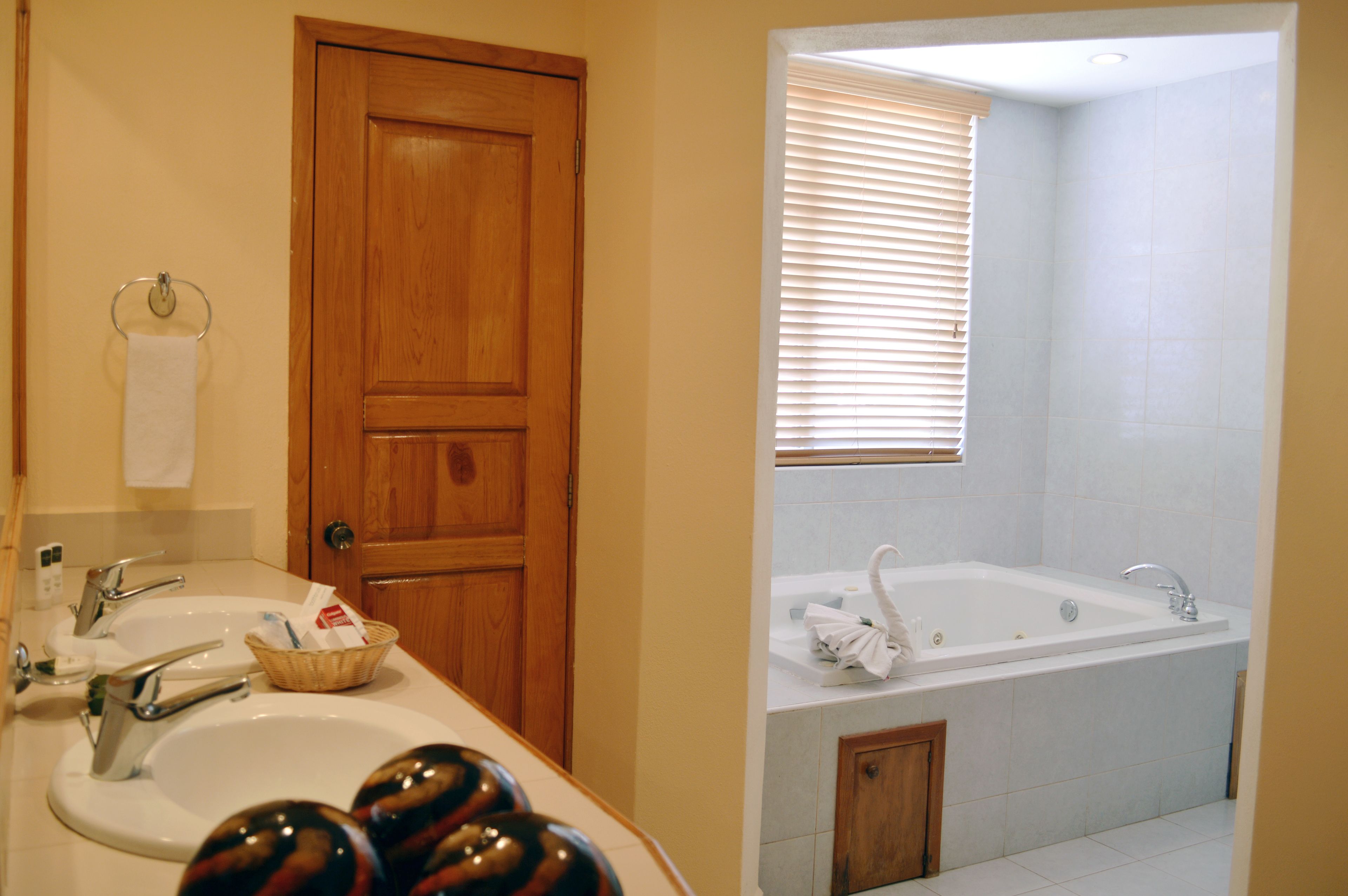 A bathroom with a wooden door, two sinks, a bathtub, and a window with blinds.