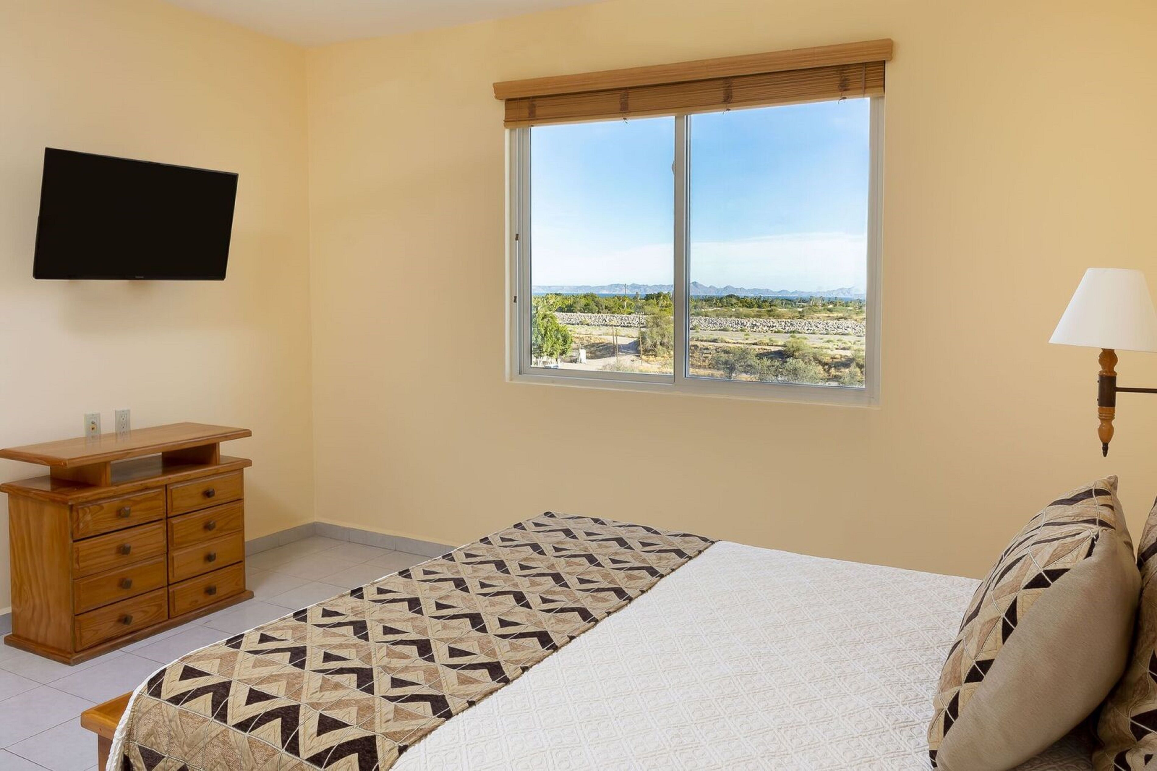 A bedroom with a bed, a wooden dresser, a flat-screen TV, a window with a view of a landscape, and a bedside lamp.