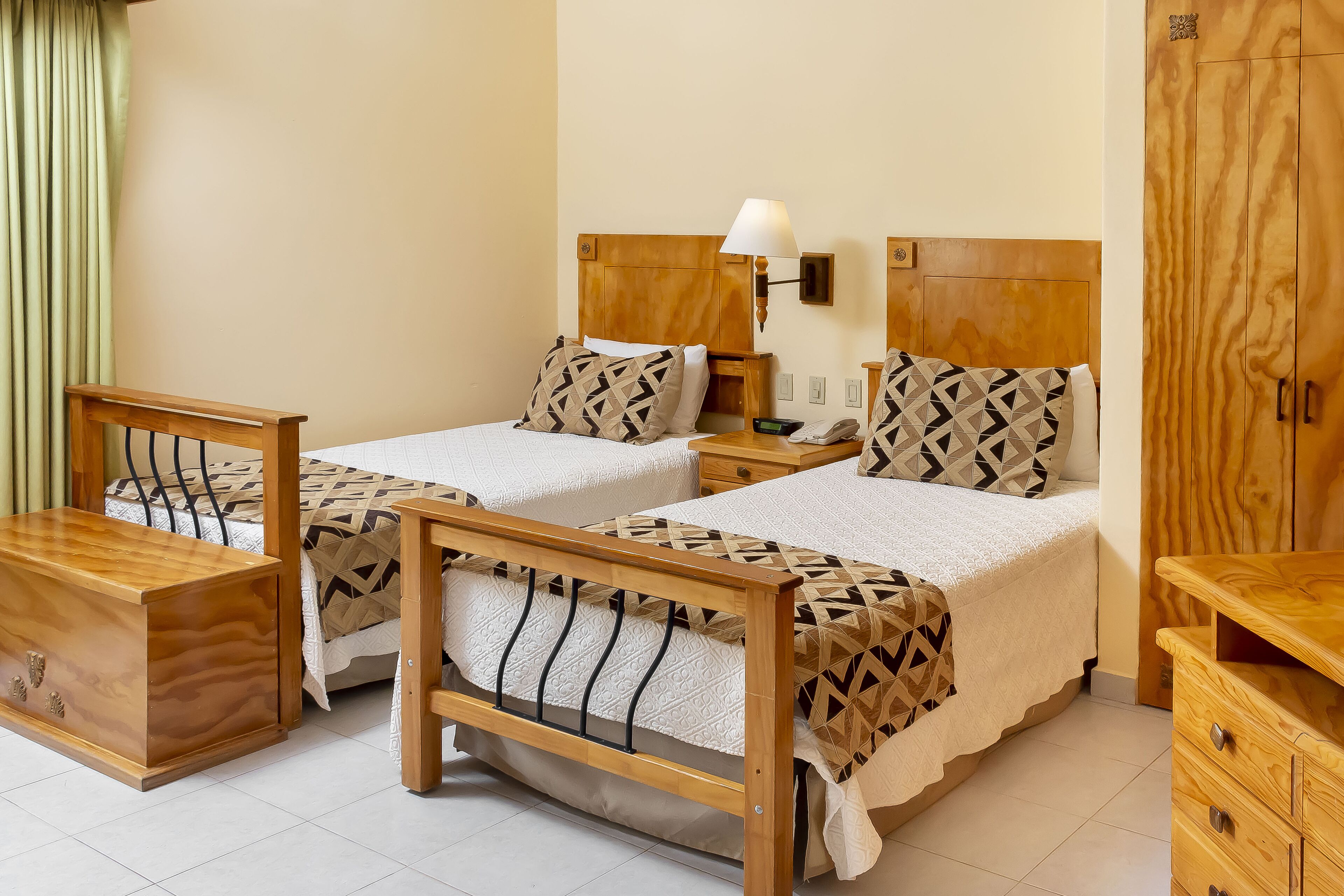 A hotel room with two beds, wooden headboards, a nightstand, a lamp, a wardrobe, and a wooden chest of drawers.