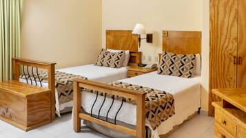A hotel room with two beds, wooden headboards, a nightstand, a lamp, a wardrobe, and a wooden chest of drawers.