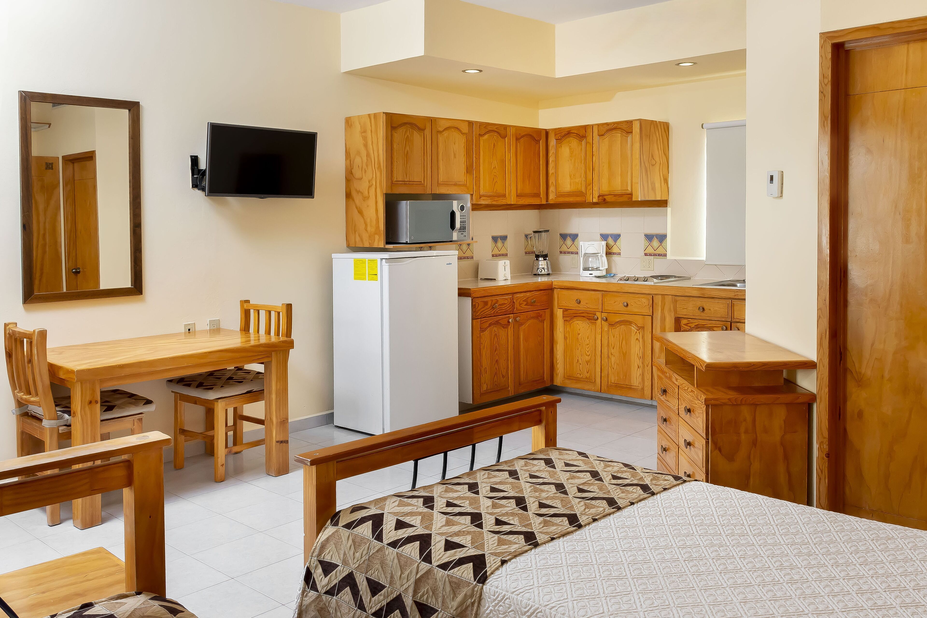 A compact room with a bed, a small dining table with chairs, a kitchenette with wooden cabinets, a refrigerator, a microwave, and a television mounted on the wall.