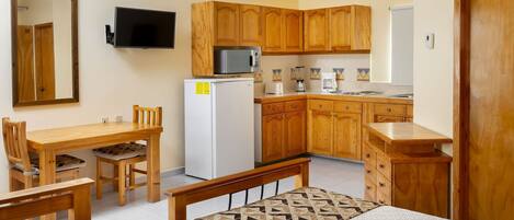 A compact room with a bed, a small dining table with chairs, a kitchenette with wooden cabinets, a refrigerator, a microwave, and a television mounted on the wall.