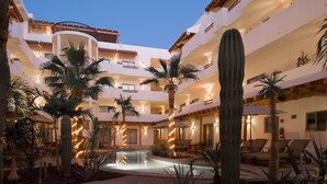 Un patio de hotel con piscina, palmeras, cactus y mobiliario exterior.