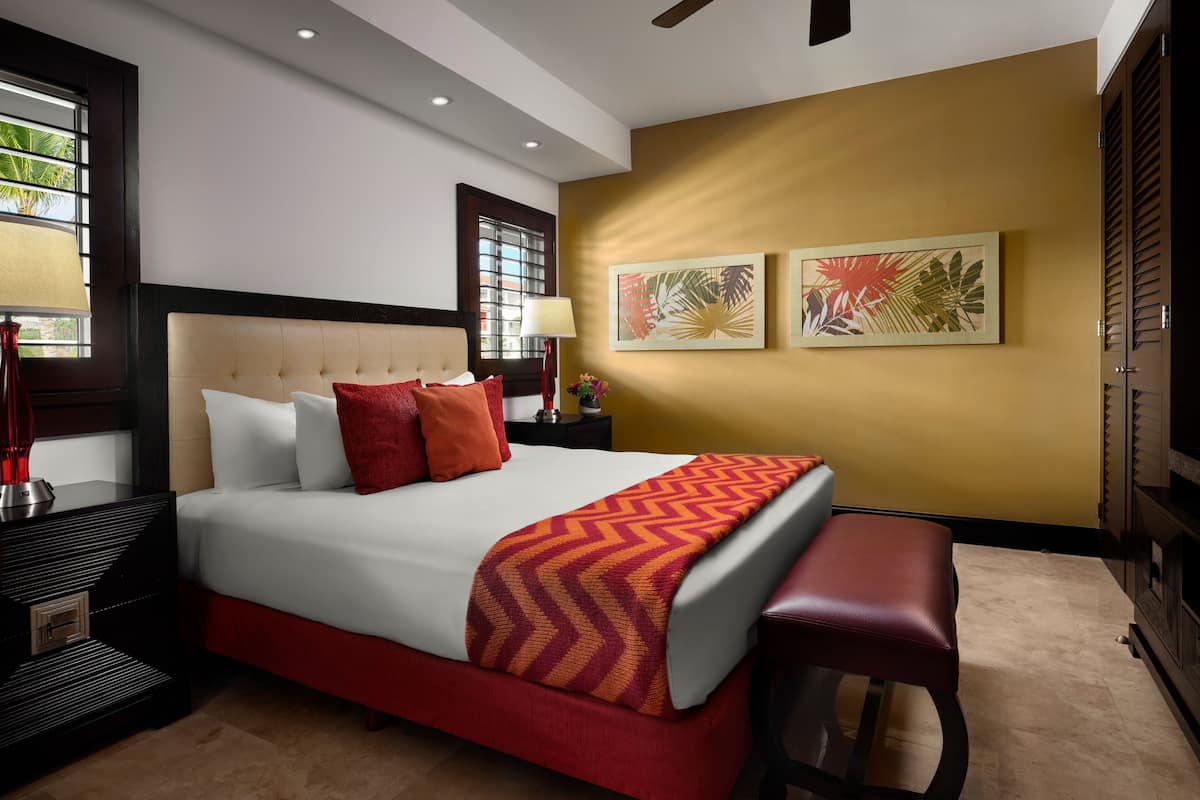 Quarto de hotel com uma cama grande, um banco, uma mesinha de cabeceira, um abajur e duas pinturas penduradas na parede.