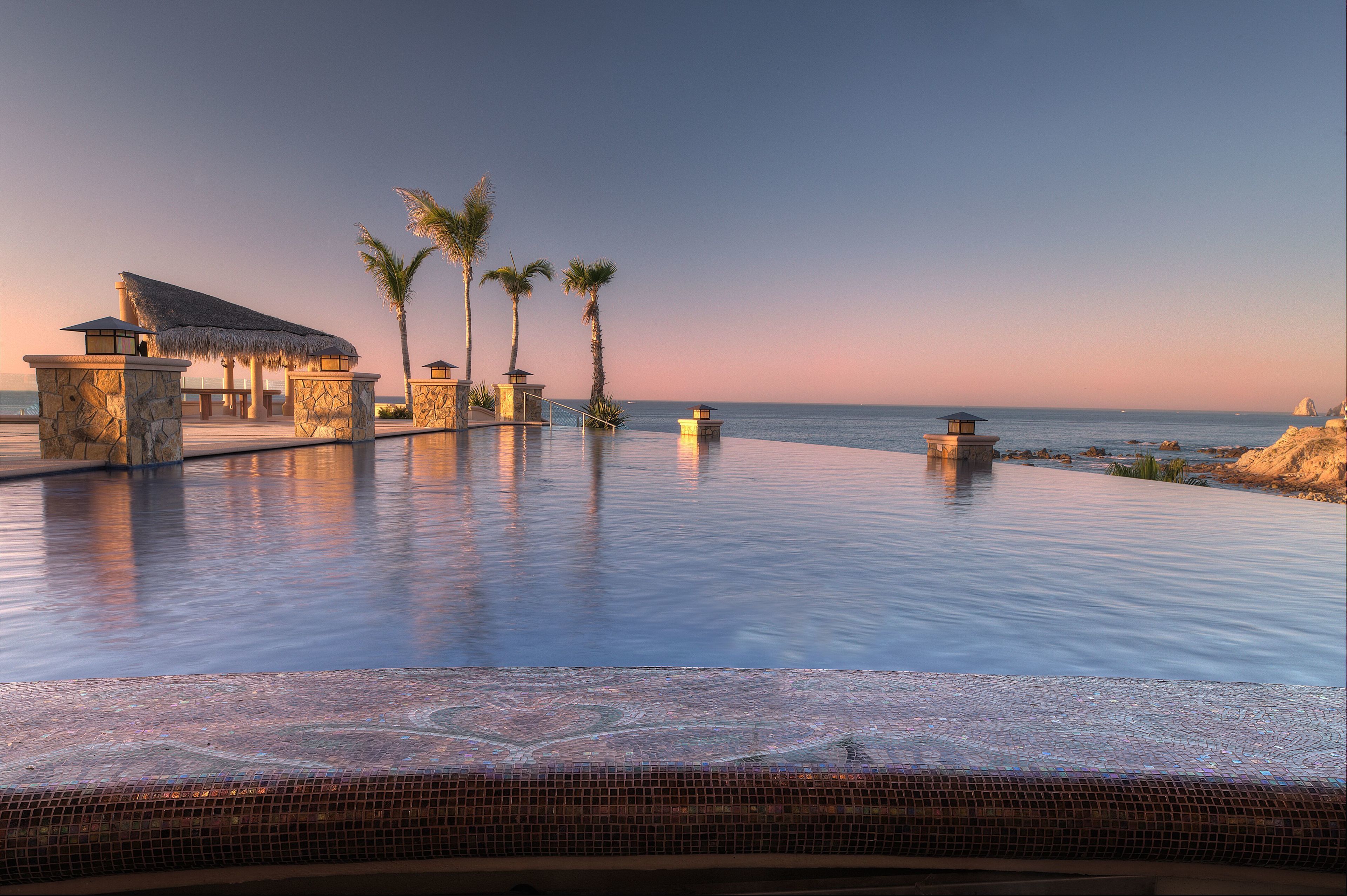 A infinity pool with a view of the ocean, palm trees, and a stone structure.