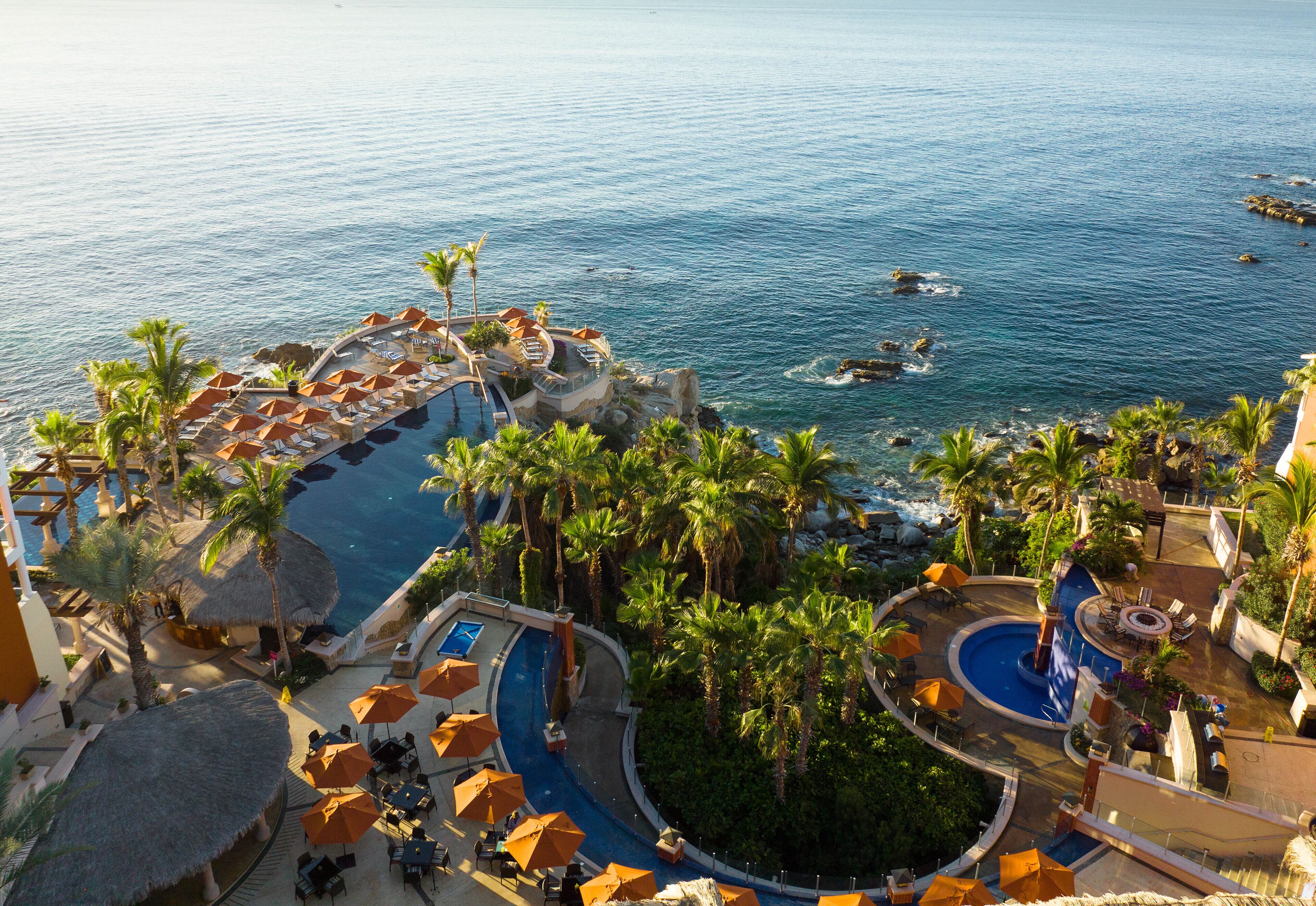 An aerial view of a resort with a large pool, palm trees, and a view of the ocean.