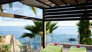 Een wellnessruimte aan zee met twee massagetafels onder een pergola, met uitzicht op de oceaan.