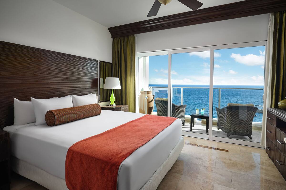 Quarto de hotel com uma cama grande, um ventilador de teto, mesa de cabeceira com abajur, vista do oceano através de uma porta de correr e uma varanda com cadeiras.
