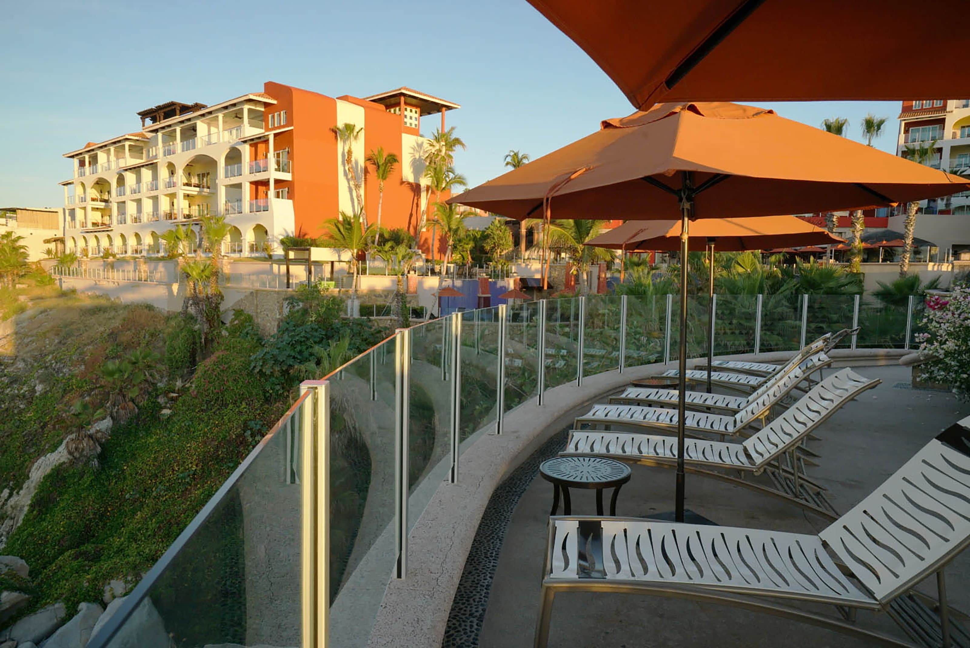 Un resort con área de piscina que cuenta con sillones de descanso y sombrillas, rodeado por una baranda de cristal y palmeras.