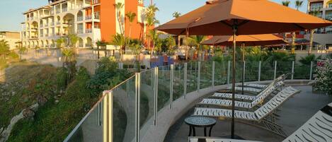 A resort with a poolside area featuring lounge chairs and umbrellas, surrounded by a glass railing and palm trees.