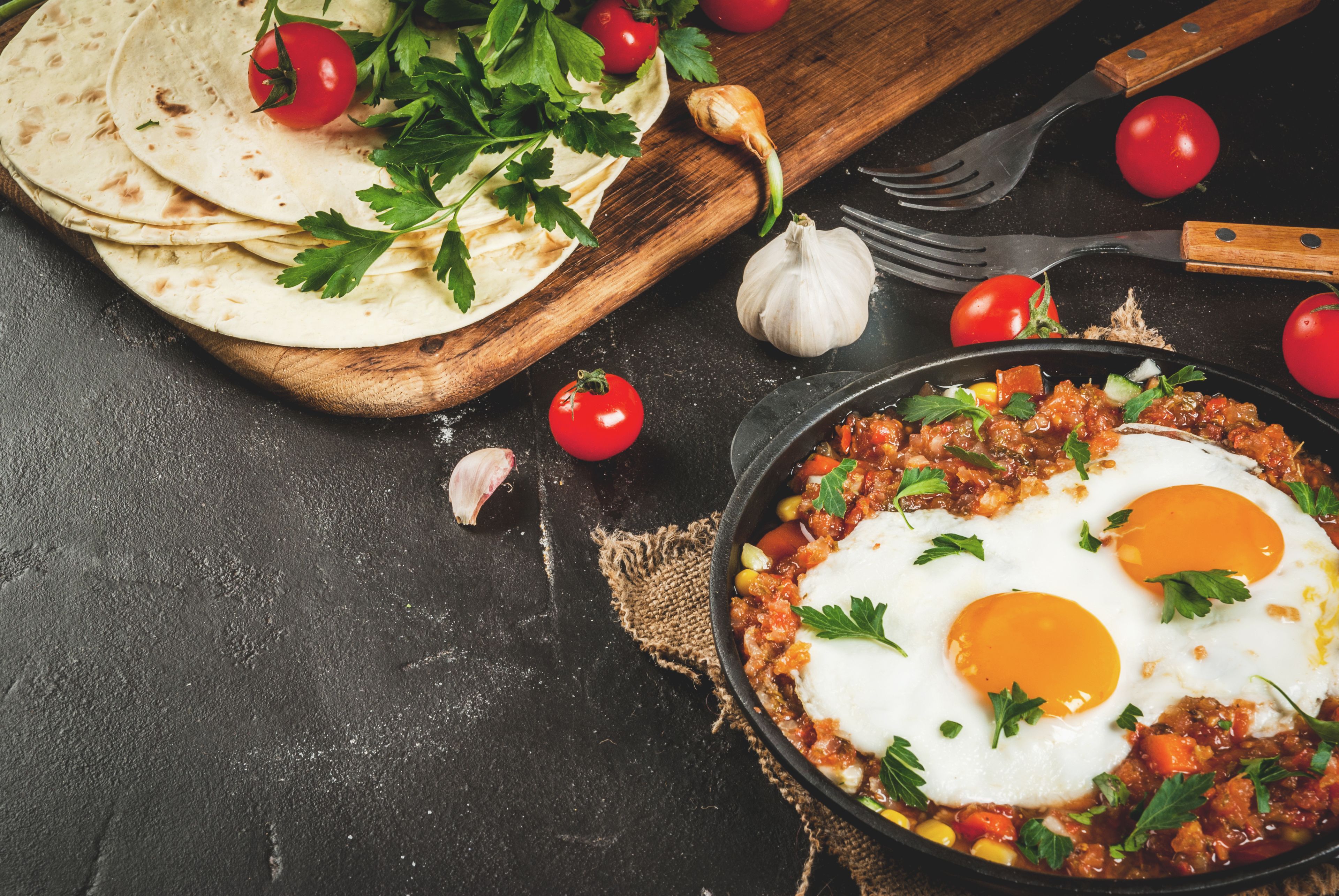 A skillet with eggs and a tomato-based dish, surrounded by fresh tomatoes, garlic, and herbs on a dark surface.