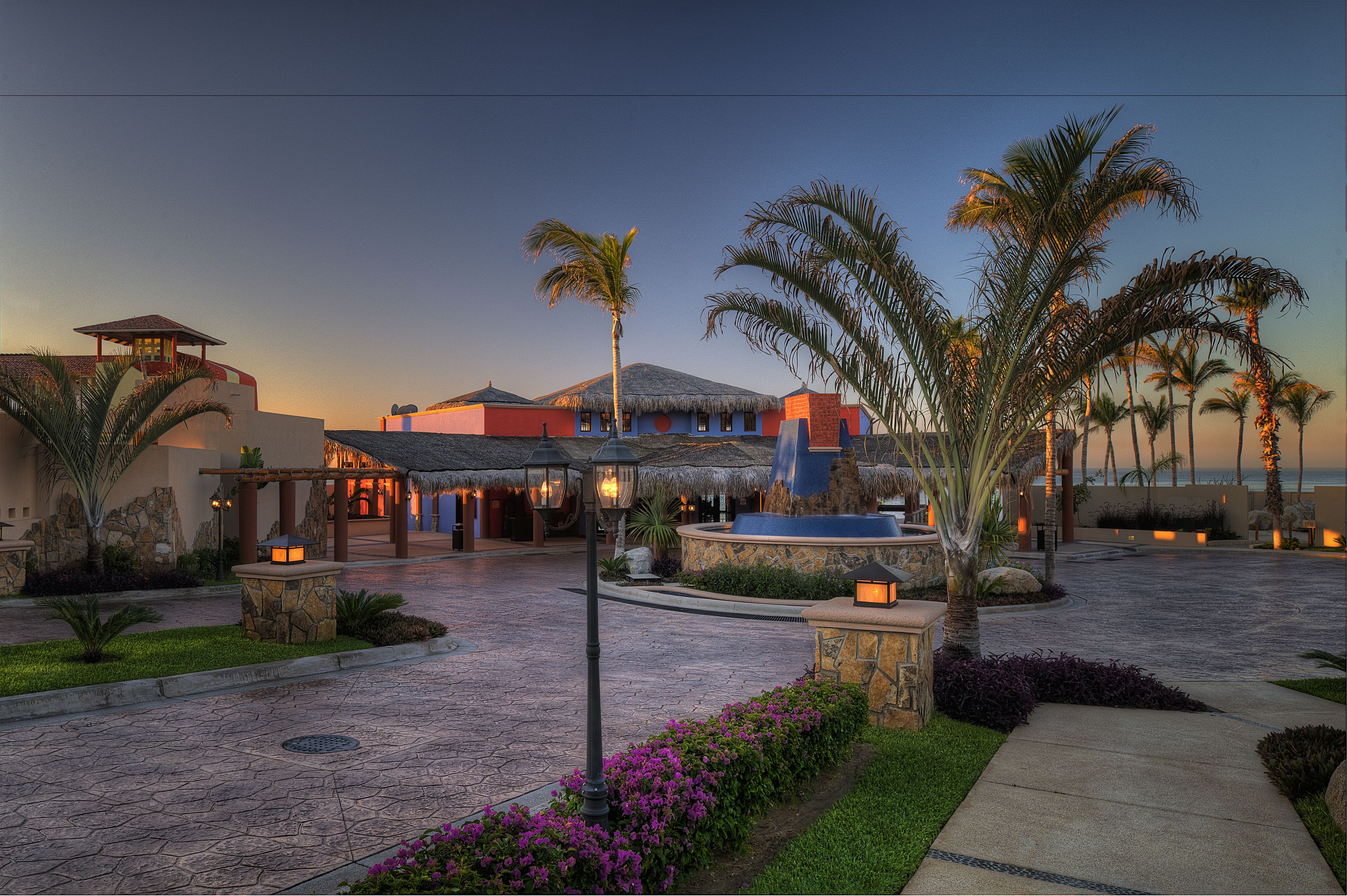 A resort courtyard with a pool, palm trees, and a thatched-roof building.
