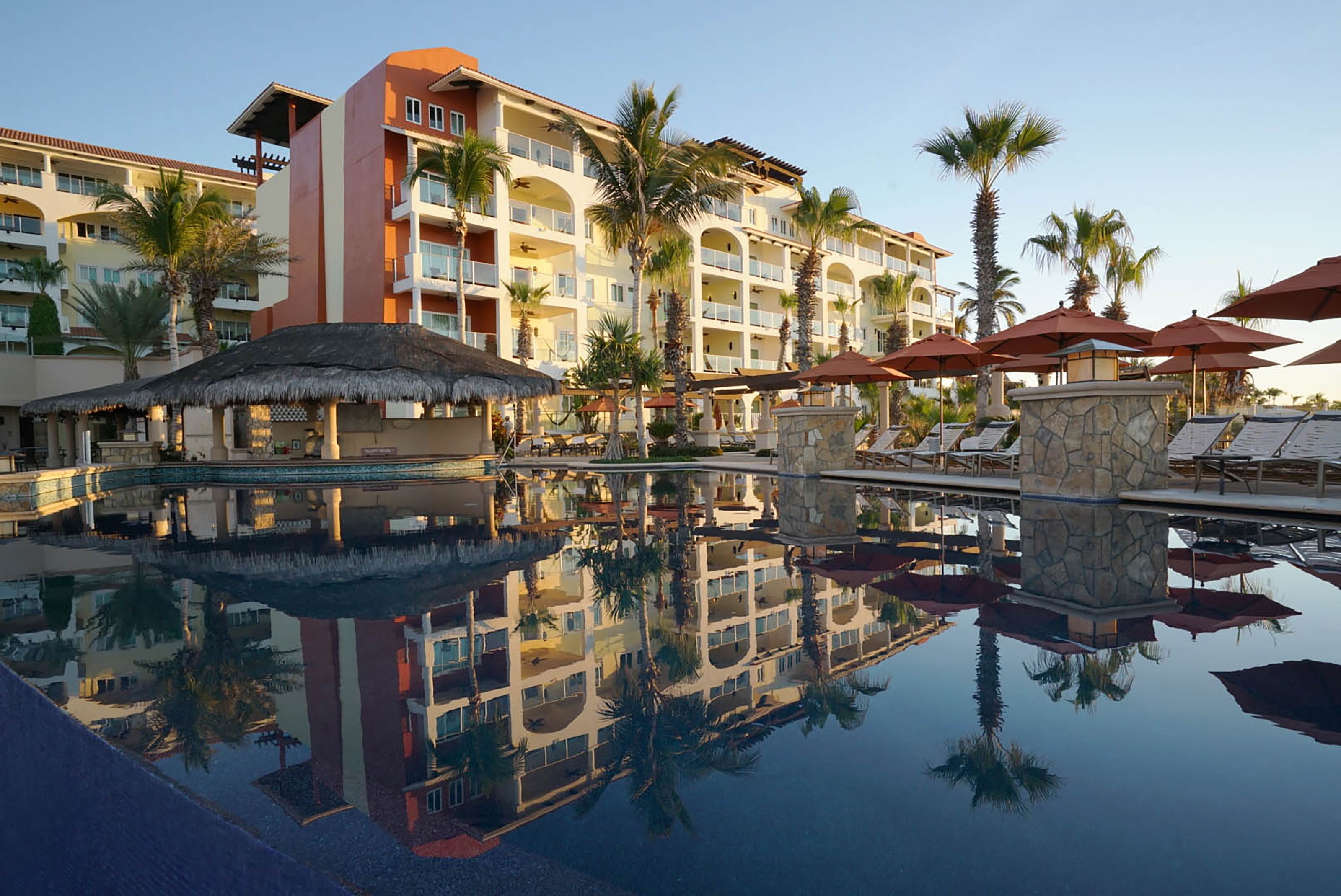 A hotel with a pool reflecting the building and palm trees, multiple balconies, and outdoor umbrellas.