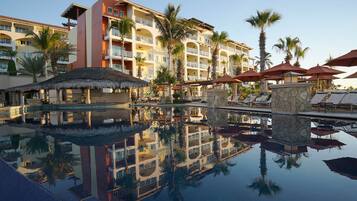 Un hotel con piscina que refleja el edificio y palmeras, varios balcones y sombrillas exteriores.