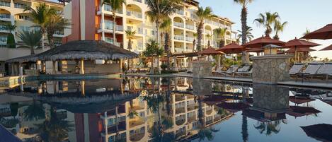 A hotel with a pool reflecting the building and palm trees, multiple balconies, and outdoor umbrellas.