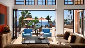 Een ruime woonkamer met een groot raam met uitzicht op de oceaan, ingericht met blauwe en bruine banken, een salontafel en twee parasols.