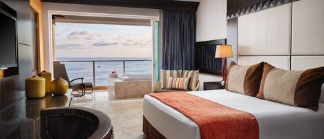 A hotel room with a large bed, a seating area with a view of the sea, a TV, and a minibar.
