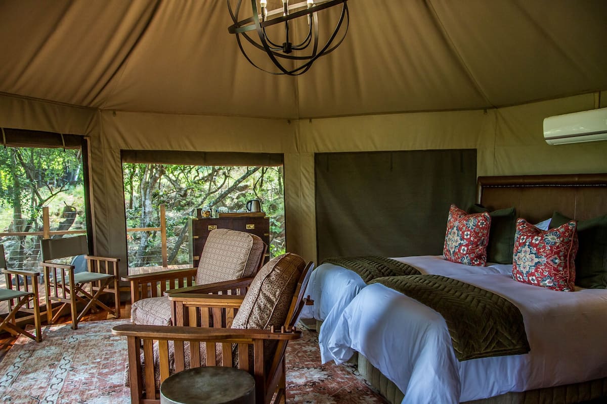 A tented accommodation with a bed, chairs, a small table, and a view of the outdoors.