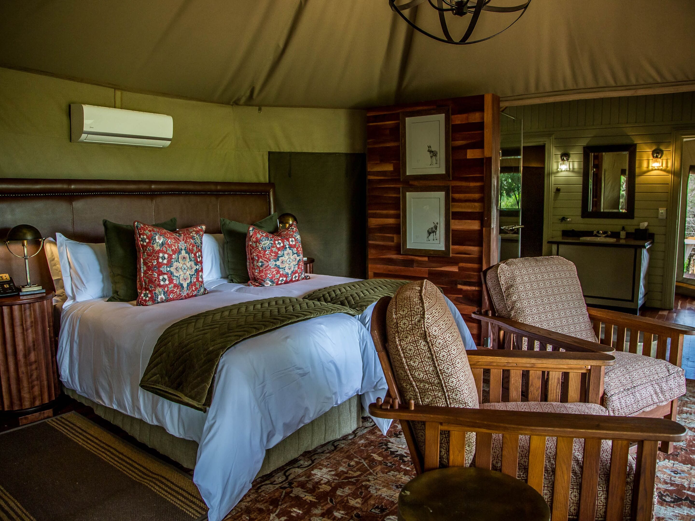 Ein Schlafzimmer mit einem großen Bett, vielen Kissen, einem Holzkopfende, einem Nachttisch mit Lampe, einem Sessel, einem Spiegel und einem kleinen Badezimmerbereich.