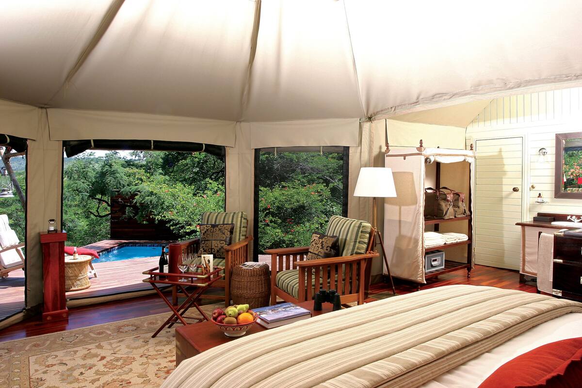 A tented room with a bed, chairs, a small table with fruit, and a view of a pool and greenery outside.