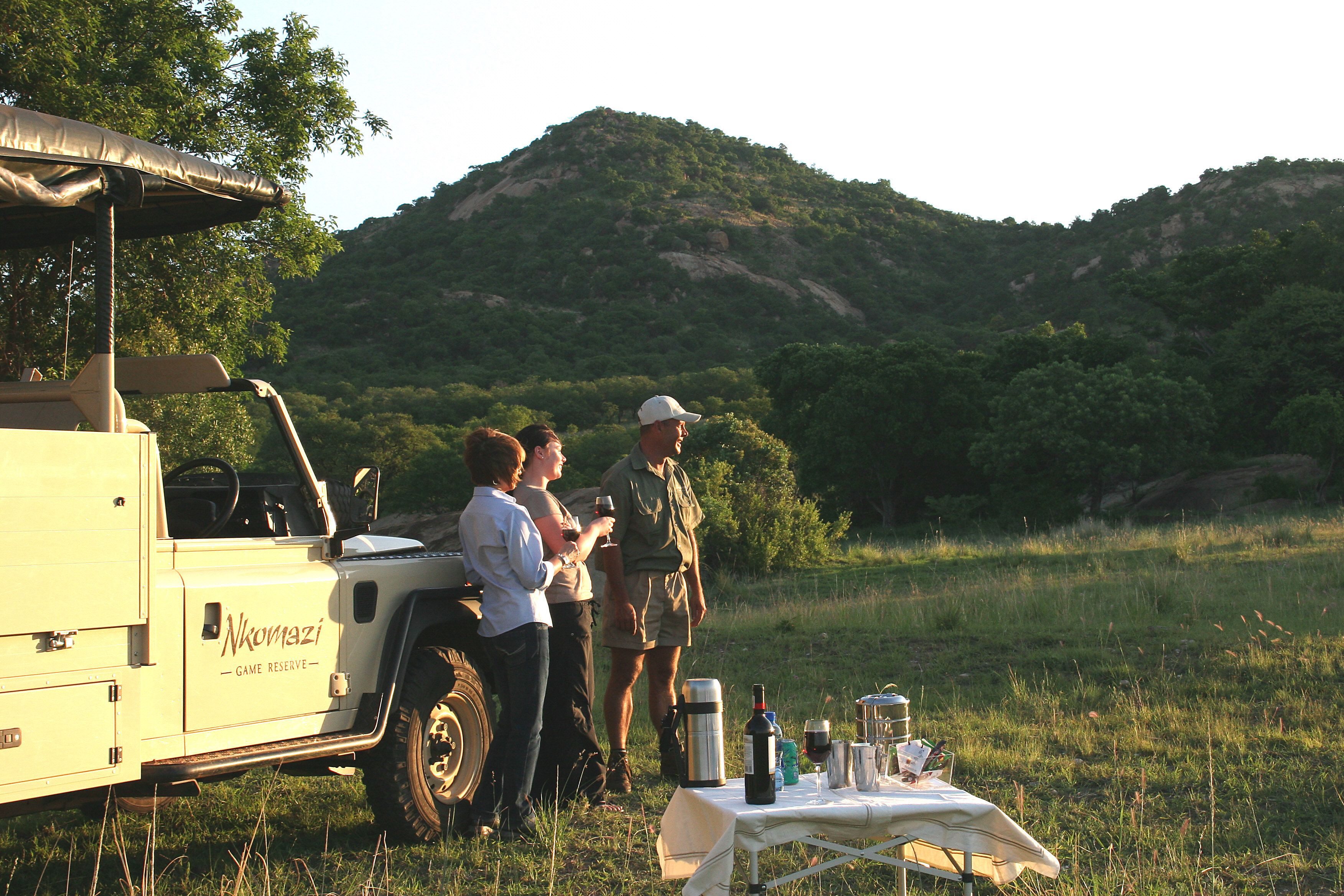Un grupo de personas de pie junto a una mesa con una tienda de campaña y un vehículo que indica "Nkowazi Game Reserve" en una zona con césped y colinas al fondo.