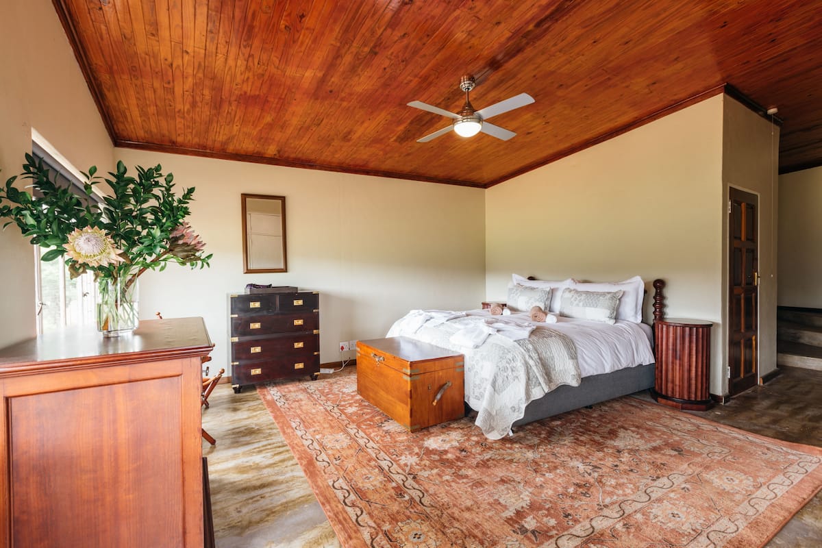 A bedroom with a wooden ceiling, a bed with white bedding, a wooden chest, a dresser, and a vase with flowers.