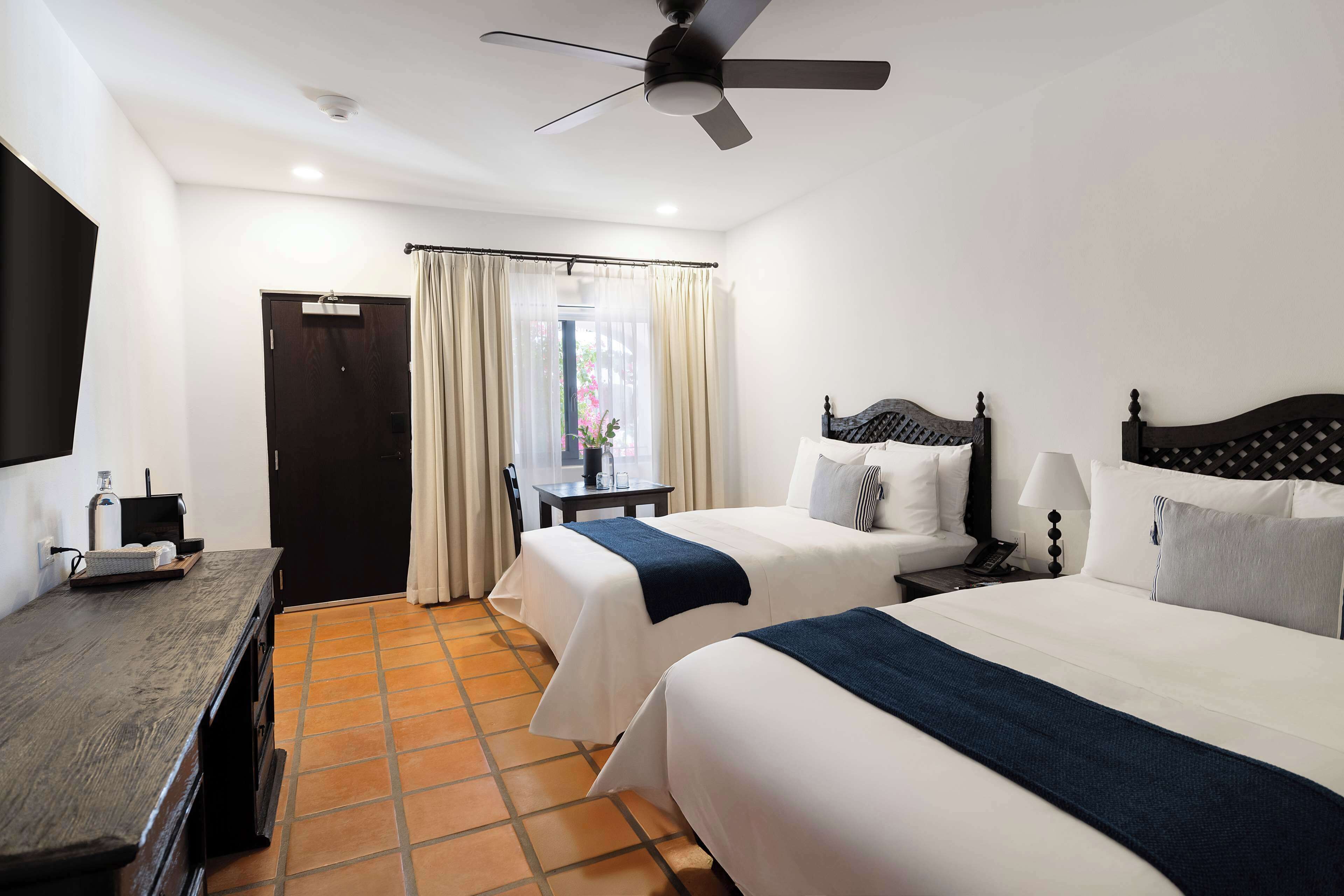 Habitación de hotel con dos camas, un escritorio, una silla, un ventilador de techo y una ventana con cortinas.
